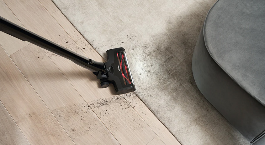 Miele Bodenbürste mit rot-schwarzer Bürstenrolle saugt Schmutz am Übergang von hellem Holzboden zu einem hellen Teppich neben einem grauen Sofa.