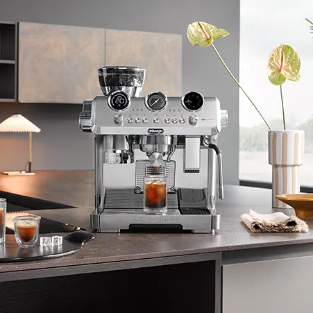 Eine silberne De'Longhi Kaffeemaschine auf einer Küchentheke mit einem Glas Kaffee. Im Hintergrund befinden sich Schränke und eine Vase mit grünen Blumen.