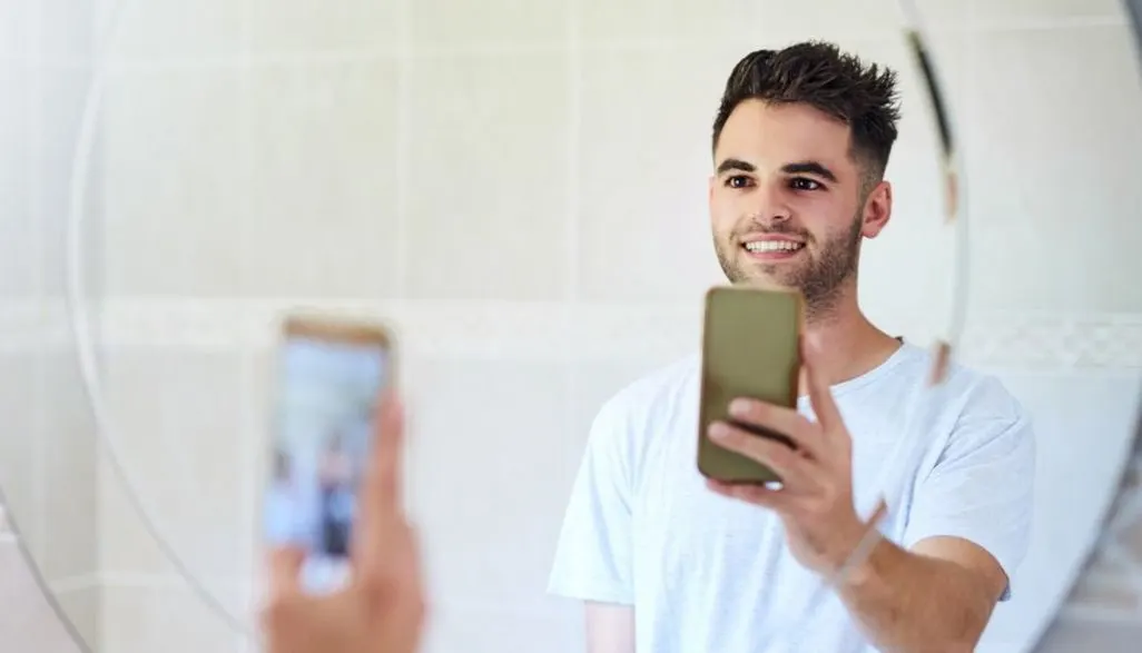 Mann macht ein Selfie mit dem Smartphone vor einem Spiegel im Badezimmer.