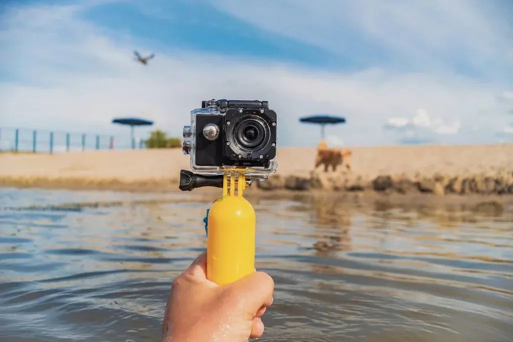 Hand mit einer Actioncam zeigt in Richtung Strandufer mit einigen Schirmen.