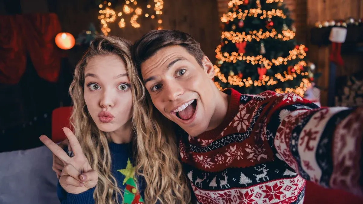 Zwei Personen in Weihnachtspullovern machen ein Selfie. Eine Person zeigt ein Friedenszeichen, die andere lächelt. Im Hintergrund ein beleuchteter Weihnachtsbaum und ein Kamin mit Strumpf.