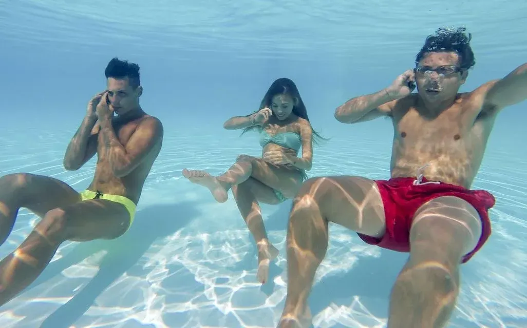 Drei Personen tauchen in klarem Wasser bei Sonnenschein und telefonieren mit einem Smartphone.