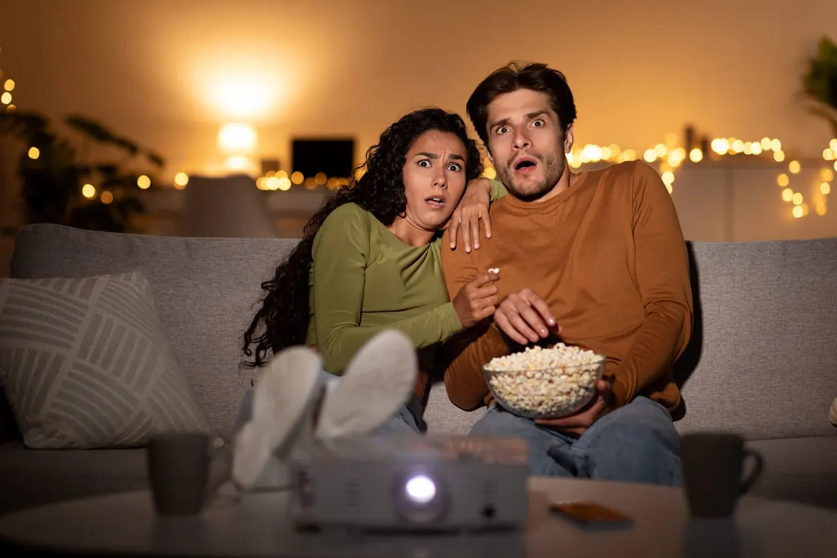 Ein junges Paar sitzt abends erschrocken auf einer grauen Couch und schaut einen Film, der von einem Beamer auf einem Tisch davor projiziert wird. Beide haben die Münder weit geöffnet und schauen mit großen Augen in Richtung Leinwand, während der Mann eine Schüssel Popcorn hält.