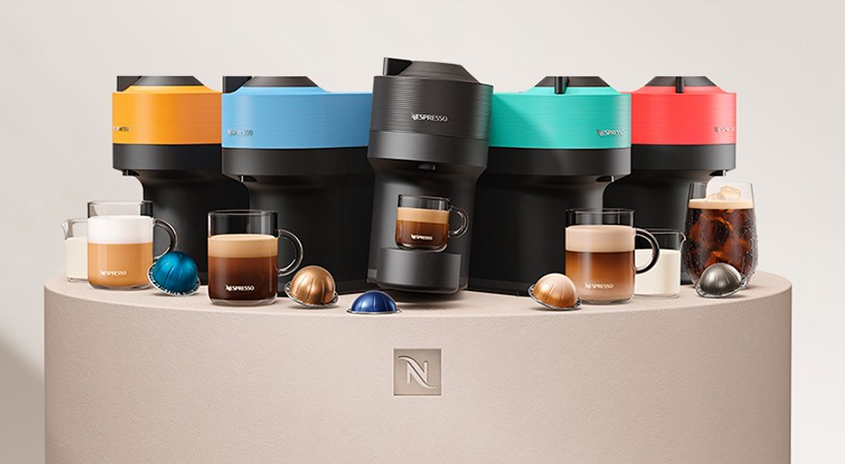 Bunte Nespresso Vertuo Pop Maschine auf Podest mit Kapseln und Kaffeebechern