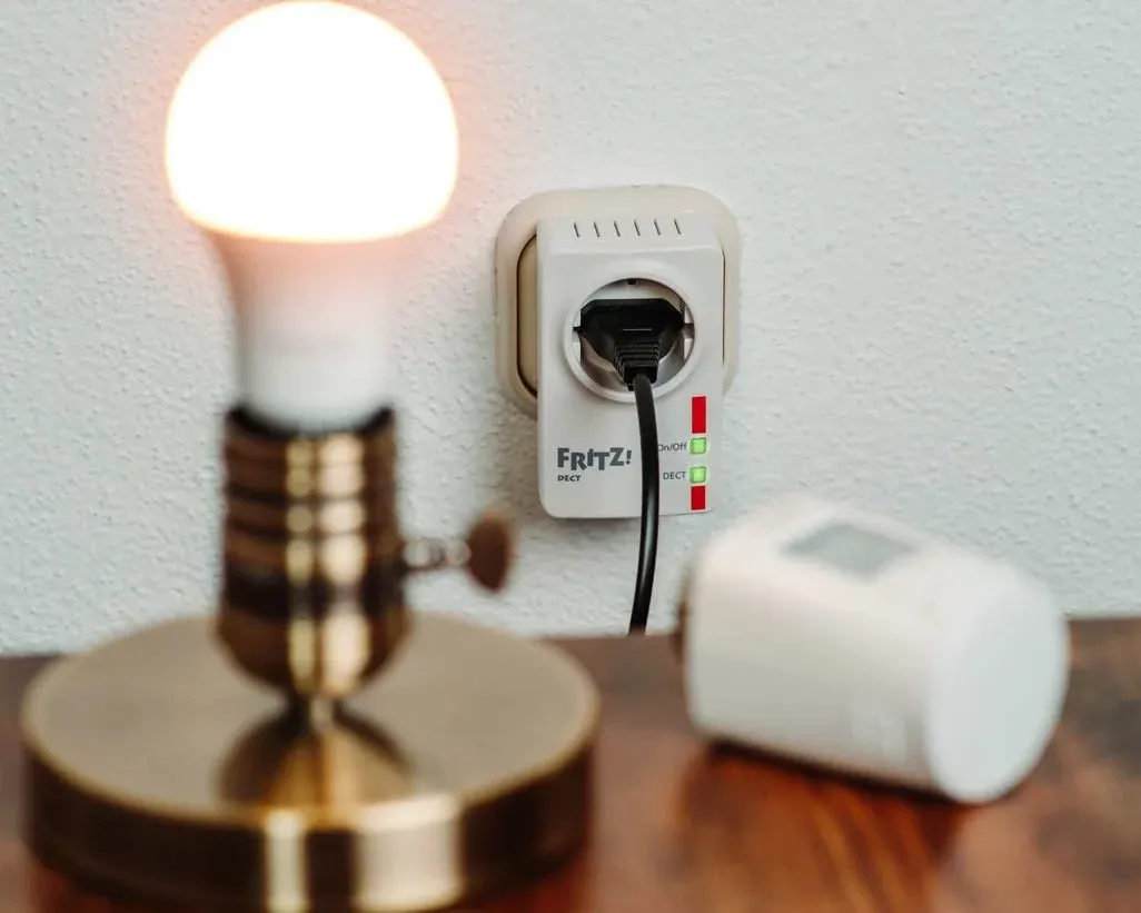 Leuchtende Glühbirne auf Messingfuß, weißer Fritz!DECT Smart Plug mit grün leuchtenden LEDs in der Wand und ein weißes Gerät auf Holz.