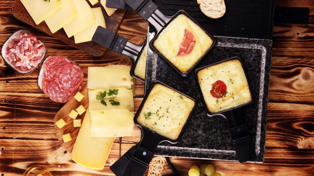 Drei gefuellte Raclette-Pfannen mit Kaese neben Aufschnitt und Kaesewuerfeln