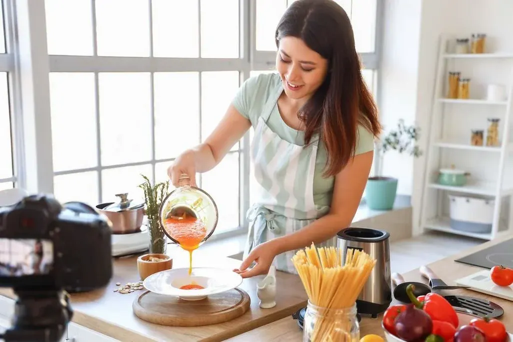 Person in moderner Küche filmt ein Kochvideo, während sie Suppe auf einen Teller gießt; im Vordergrund stehen Mixer, Gemüse und Pasta.