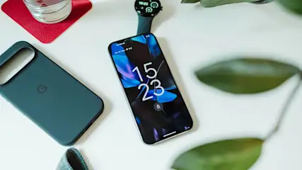 Google Pixel 9 Pro Smartphone liegt auf einem Tisch neben einer schwarzen Smartwatch, einer Google-Handyhülle, einem Glas Wasser und gruenen Blättern.