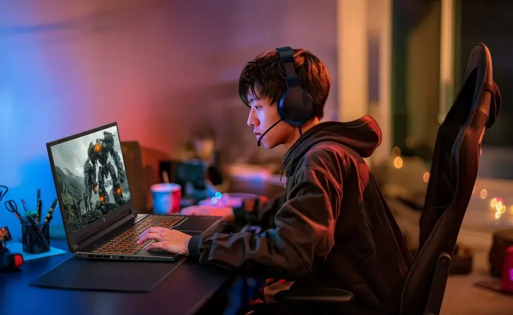 Gamer spielt an Laptop mit leuchtender Tastatur und Headset. Bildschirm zeigt Roboterspiel. Raum ist mit blauem und rotem Licht beleuchtet.