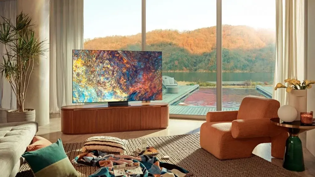 Ein Wohnzimmer mit einem großen Fenster mit Blick auf einen See und Berge, einem Fernseher und Möbeln.
