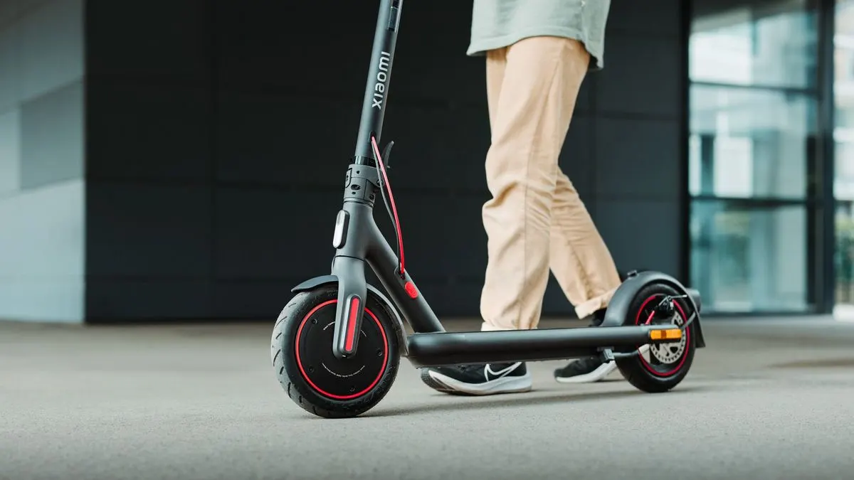 Schwarzer Xiaomi Elektroroller mit roten Akzenten auf grauem Pflaster wird von einer Person in heller Hose geschoben.