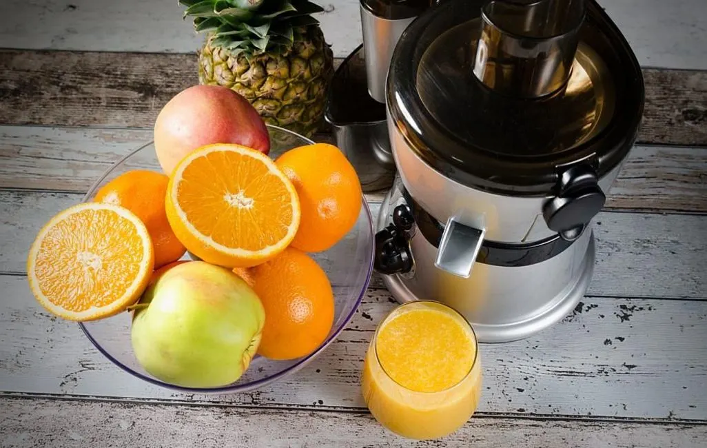 Obstschale mit Orangen, Apfel und Ananas neben einem Entsafter und einem Glas Saft