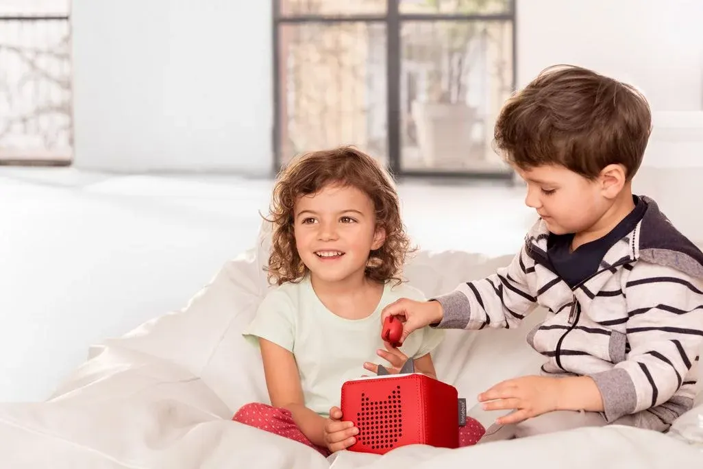 Zwei Kinder sitzen auf einem weißen Sitzsack. Ein Mädchen hält eine rote Toniebox, ein Junge setzt eine rote Figur darauf. Im Hintergrund ein Fenster mit Pflanzen.