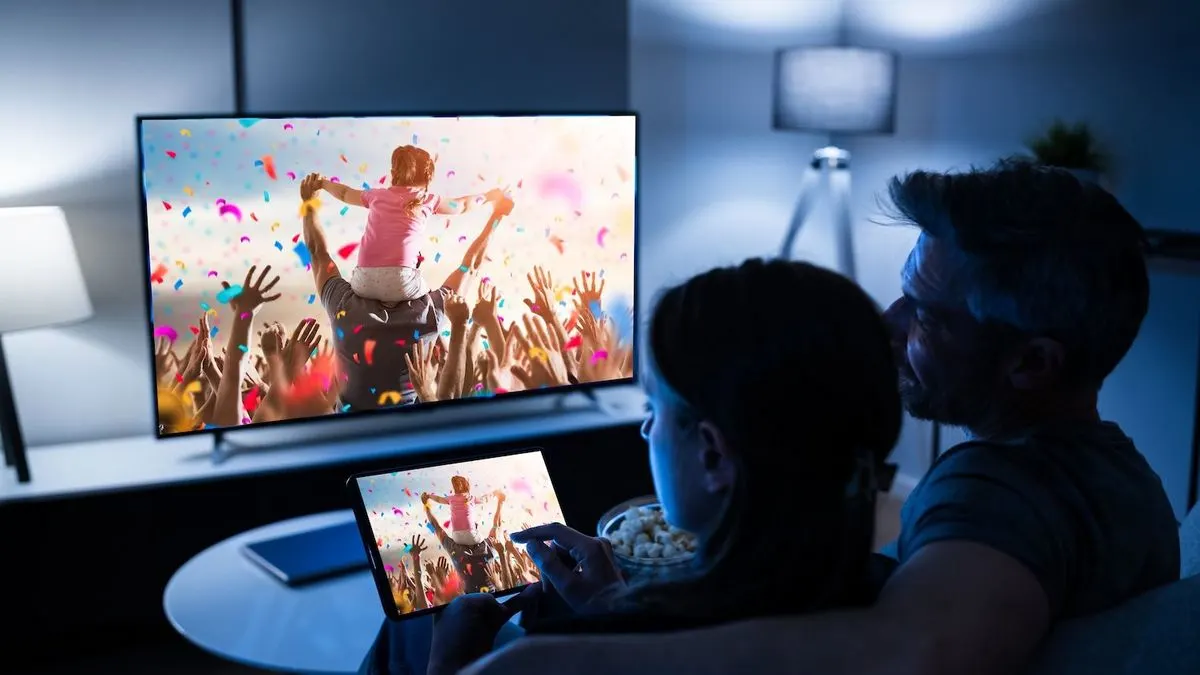 Zwei Personen sehen im dunklen Raum fern. Eine Person hält Tablet und Popcorn. TV und Tablet zeigen ein Kind auf Schultern mit Konfetti.