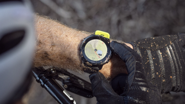Ein Arm mit einer schwarzen Uhr mit gelbem Detail wird von einer Hand mit schwarzem Handschuh gehalten. Fahrradlenker sichtbar.