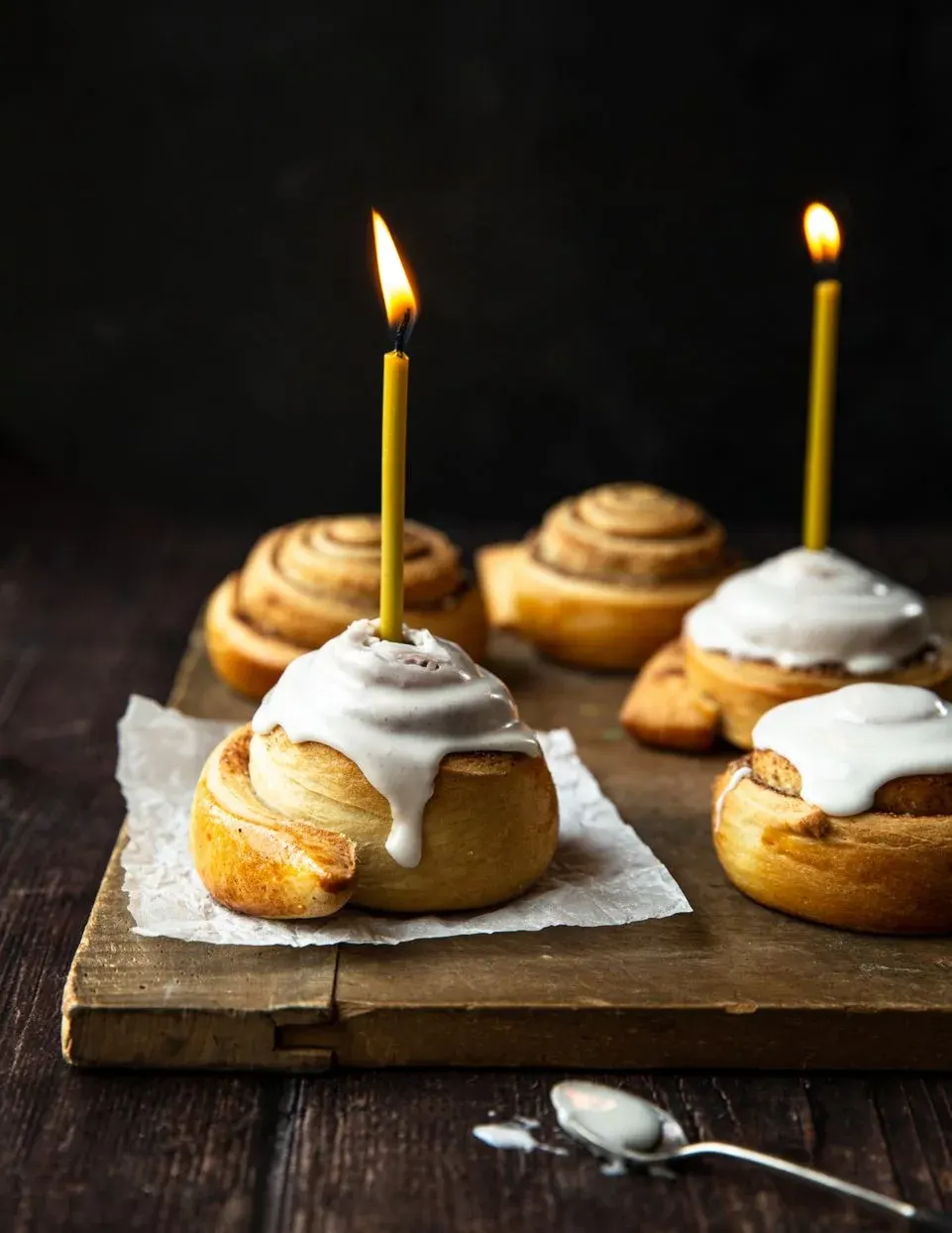 Mehrere Zimtschnecken mit weißem Zuckerguss und brennenden gelben Kerzen auf einem Holzbrett. Ein Löffel liegt daneben.