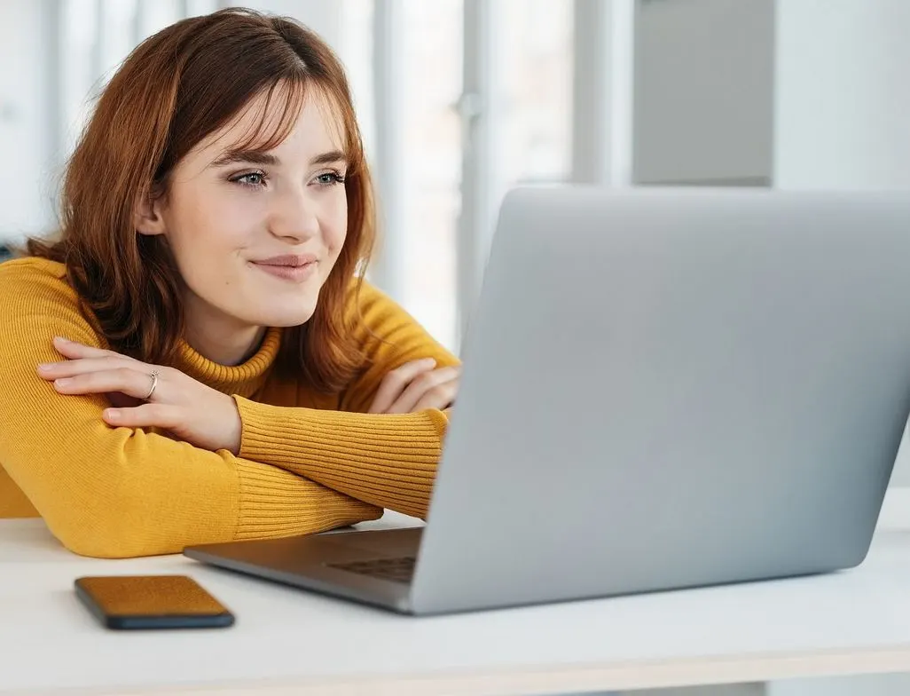 Laechelnde Frau in gelbem Pullover arbeitet entspannt am silbernen Laptop