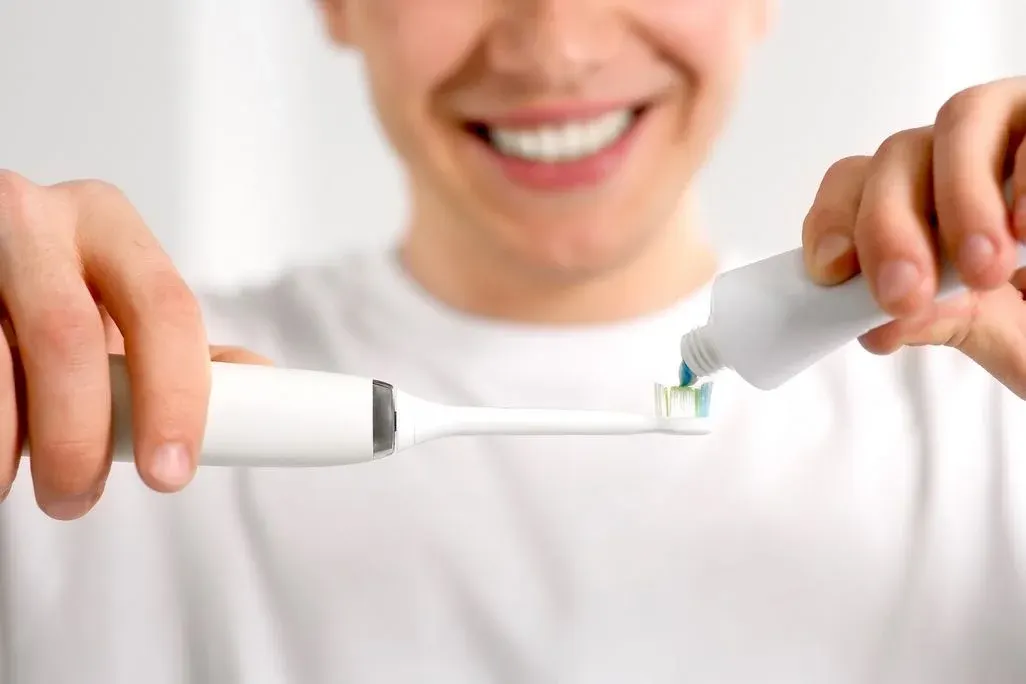 Lachende Person in weißem T‑Shirt hält eine elektrische Zahnbürste und trägt mit der anderen Hand blaue Zahnpasta aus einer Tube auf den Bürstenkopf auf.