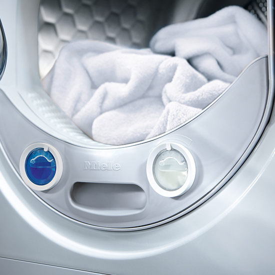 Nahaufnahme einer silbernen Waschmaschine mit weißer Wäsche im Inneren. Zwei Dosierschalter, einer blau, einer klar, befinden sich unterhalb der Öffnung.