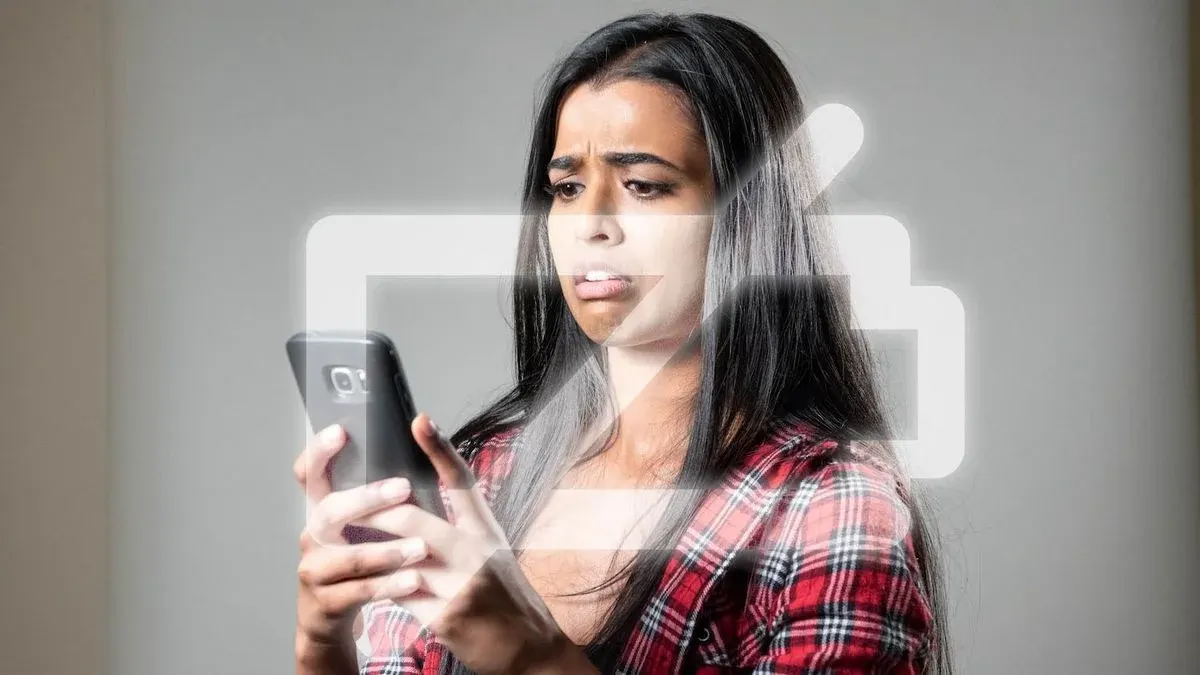 Eine Person in rotem Karohemd blickt besorgt auf ein schwarzes Smartphone. Ein leuchtendes Batteriesymbol überlagert das Bild vor grauem Hintergrund.