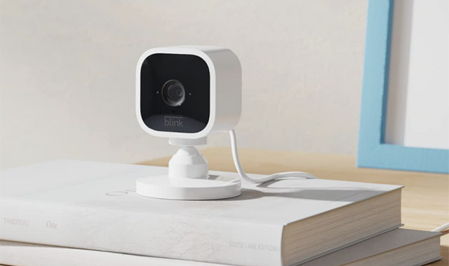 Eine weiße Überwachungskamera steht auf einem Stapel weißer Bücher vor einem blauen Bilderrahmen.