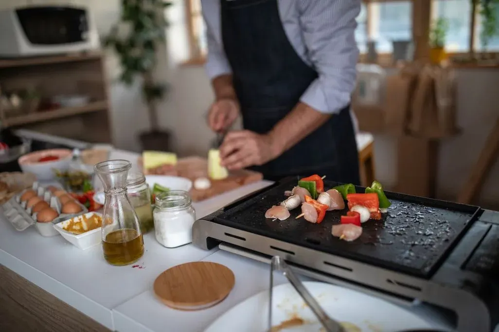 Ein Mann bereitet in einer Spieße mit Fleisch und Gemüse auf einem Elektrogrill zu, er schneidet dabei auf einem Brett etwas auf