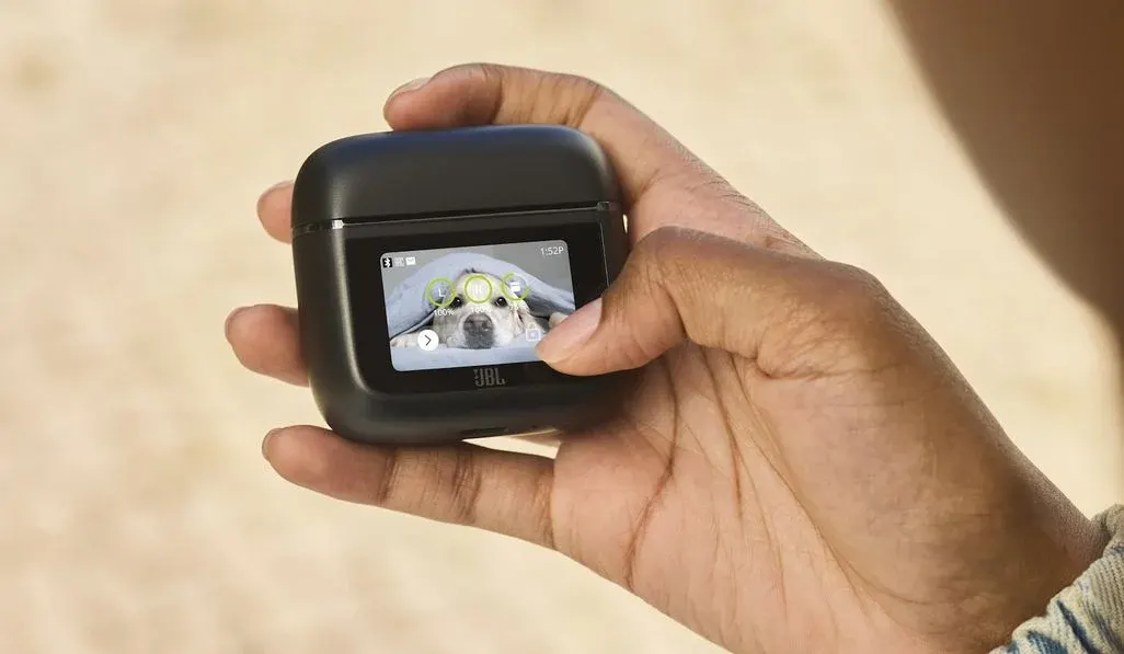 Eine Hand hält ein schwarzes JBL-Kopfhörer-Ladecase und bedient das integrierte Touch-Display. Auf dem Bildschirm ist ein Foto eines Hundes als Hintergrundbild zu sehen.
