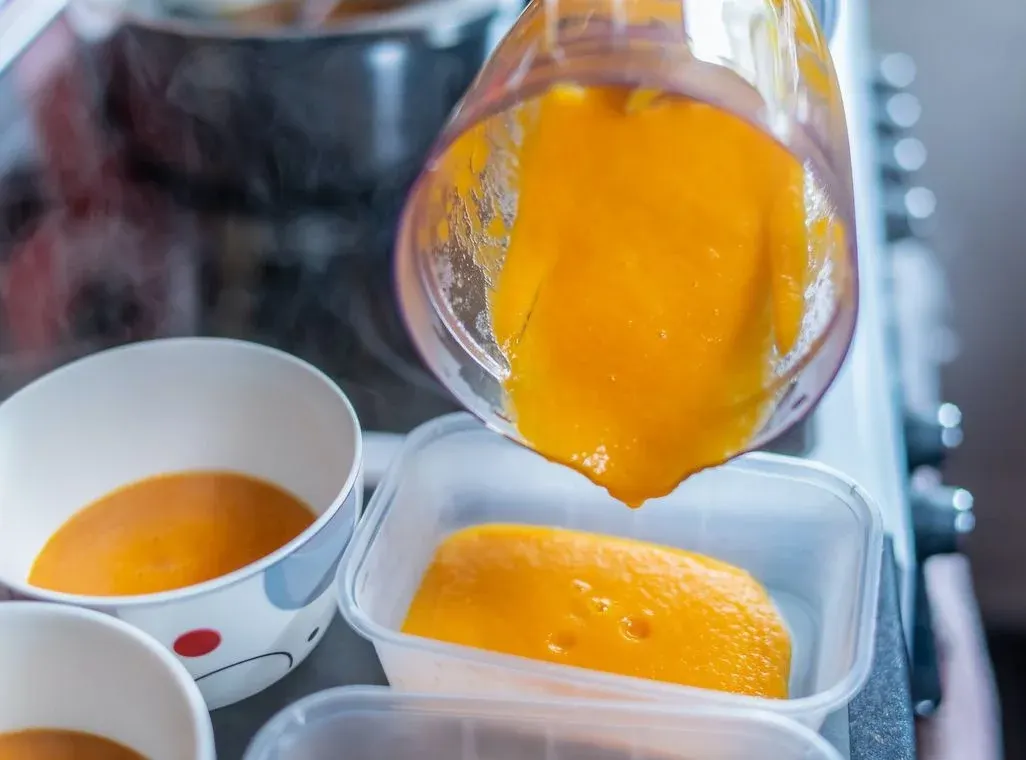 Orangenes Püree wird aus einem Mixer in transparente Behälter und weiße Schalen gefüllt. Dampf steigt im Hintergrund auf.