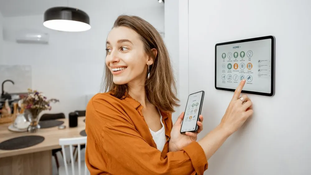 Lächelnde Frau in Orange bedient Smart-Home-Tablet und Smartphone. Im Hintergrund: moderne Küche mit Holztisch, Lampe, Klimaanlage.