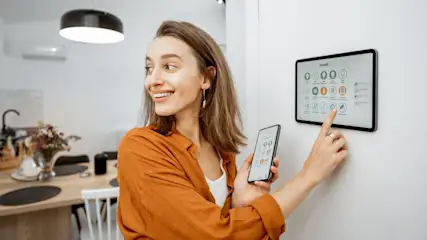 Lächelnde Frau in Orange bedient Smart-Home-Tablet und Smartphone. Im Hintergrund: moderne Küche mit Holztisch, Lampe, Klimaanlage.