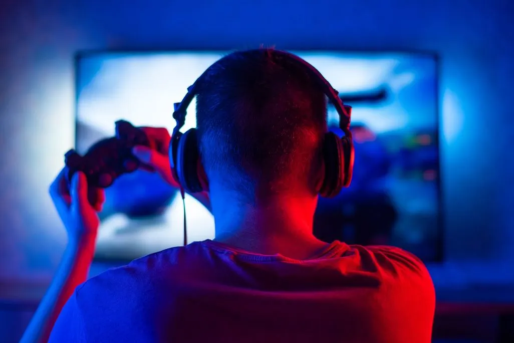 Person mit Kopfhörern sitzt vor Bildschirm und hält einen Gamecontroller, blau-rotes Licht im Raum.