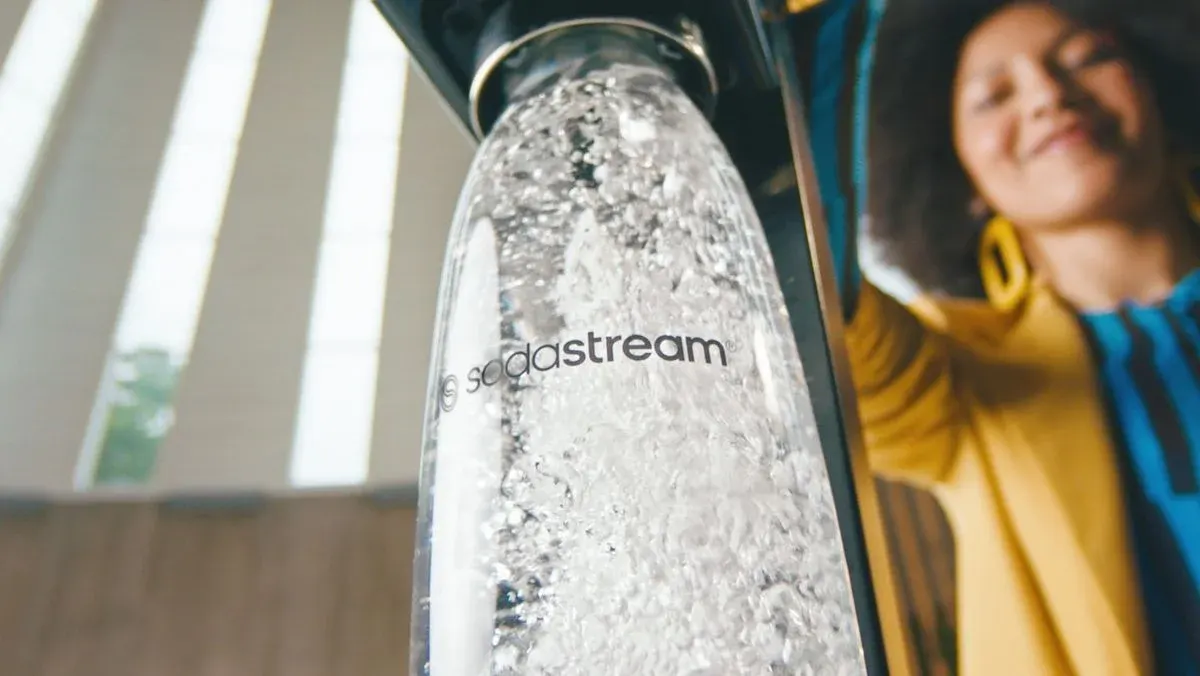 Sprudelndes Wasser in SodaStream-Flasche mit Logo an schwarzem Spender. Lächelnde Person in gelb-blauem Oberteil mit gelben Ohrringen im Hintergrund.