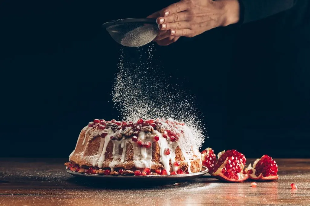 Zucker wird mit einem Sieb über einen Kuchen mit Zuckerguss und Granatapfelkernen gestäubt.