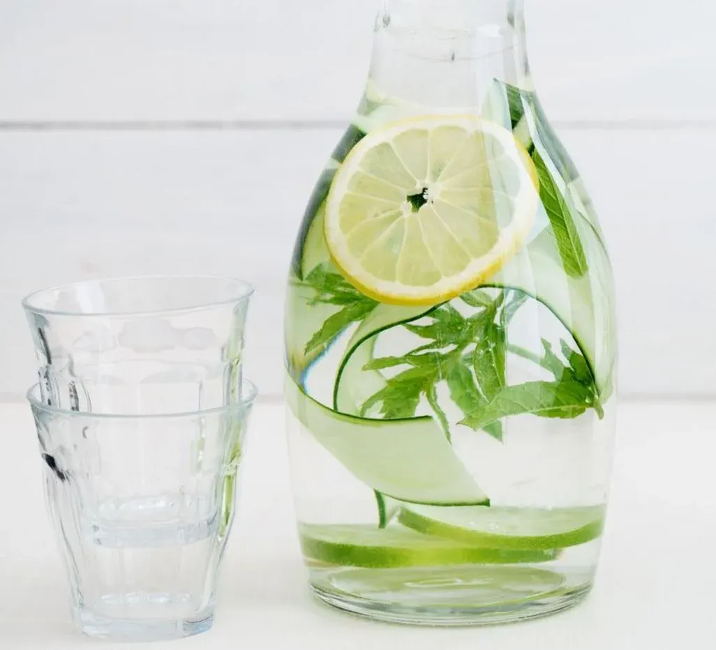  Glaskaraffe mit Wasser, in dem Zitronenscheiben, Gurkenstreifen und frische grüne Kräuter schwimmen, daneben zwei ineinander gestapelte leere Trinkgläser auf einer weißen Oberfläche.