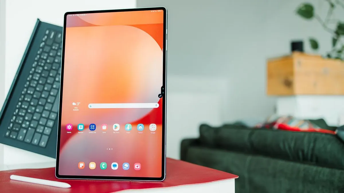 Vertikales Tablet mit orangefarbenem Bildschirm und Apps auf roter Oberfläche. Davor ein weißer Stift, links eine schwarze Tastatur. Hintergrund: grünes Sofa, Pflanze.