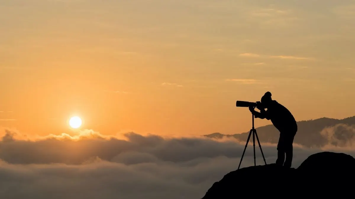 Silhouette einer Person mit Stativ, Kamera und großem Objektiv auf einem Berg bei Sonnenaufgang über einer Wolkendecke und Bergen.