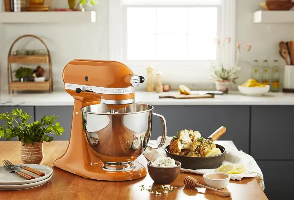 Orangefarbene Küchenmaschine mit Edelstahlschüssel auf einer Arbeitsplatte, daneben Teller, Schüssel mit Zutaten und Pfanne mit gebackenem Blumenkohl in heller Küche.
