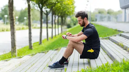 Ein Mann in schwarzer Sportkleidung sitzt auf grauen Stufen und bedient einen blauen Fitness-Tracker. Im Hintergrund sind Bäume und grüne Wiesen.