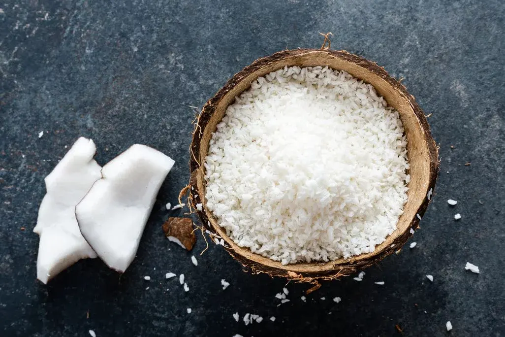 Kokosnussschale gefüllt mit geraspelter Kokosnuss, daneben Kokosfleischstücke und Schalenreste auf dunklem Grund.