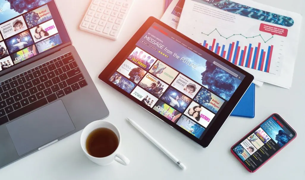 Auf einem Schreibtisch liegen ein Laptop, ein Tablet und ein Smartphone mit Streamingdienst