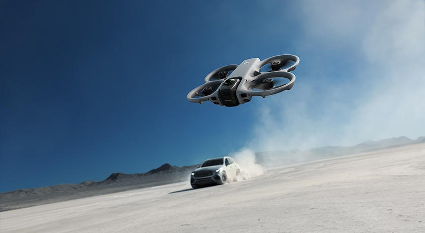  Drohne fliegt über trockene, staubige Landschaft, im Hintergrund verfolgt ein Auto die Szene und wirbelt Staub auf, blauer Himmel über Wüstenkulisse.