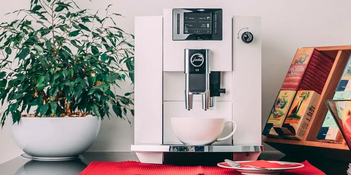 Weiße Jura E6 Kaffeemaschine mit Tasse auf roter Unterlage. Links grüne Topfpflanze, rechts Holzregal mit Teepackungen.