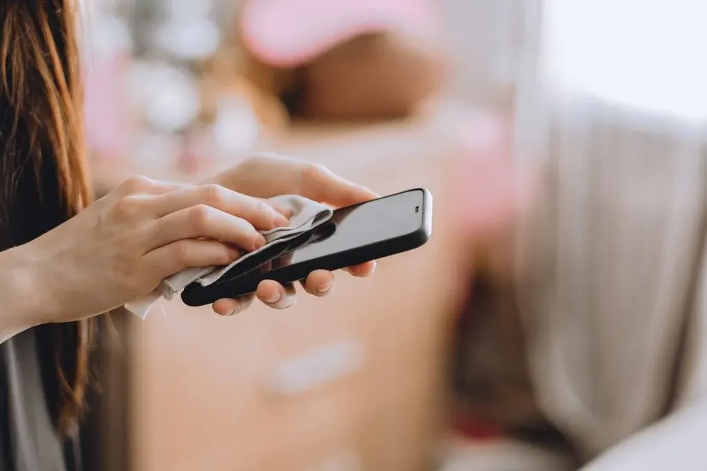 Hände wischen ein schwarzes Smartphone mit einem hellen Tuch. Unscharfer, heller Hintergrund mit rosa Akzenten.