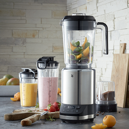 Ein Standmixer aus Edelstahl mixt Obst und Gemüse. Shakes in Bechern, Schneidebretter und Früchte sind auch zu sehen.