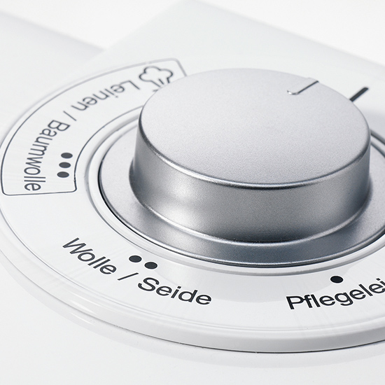 Nahaufnahme eines weißen Drehknopfes mit silberfarbenem Akzent, der auf einem Bedienfeld mit Programmsymbolen und -beschriftungen platziert ist.