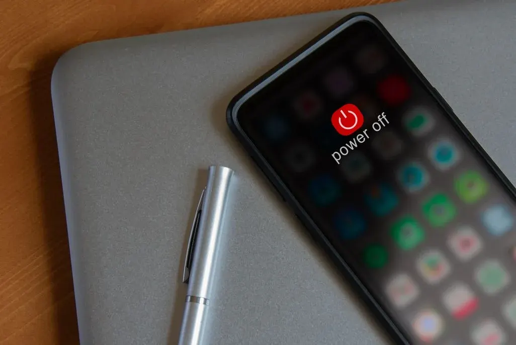 Schwarzes Smartphone mit rotem "power off" Symbol und Text, daneben ein silberner Stift auf grauem Laptop und Holztisch.