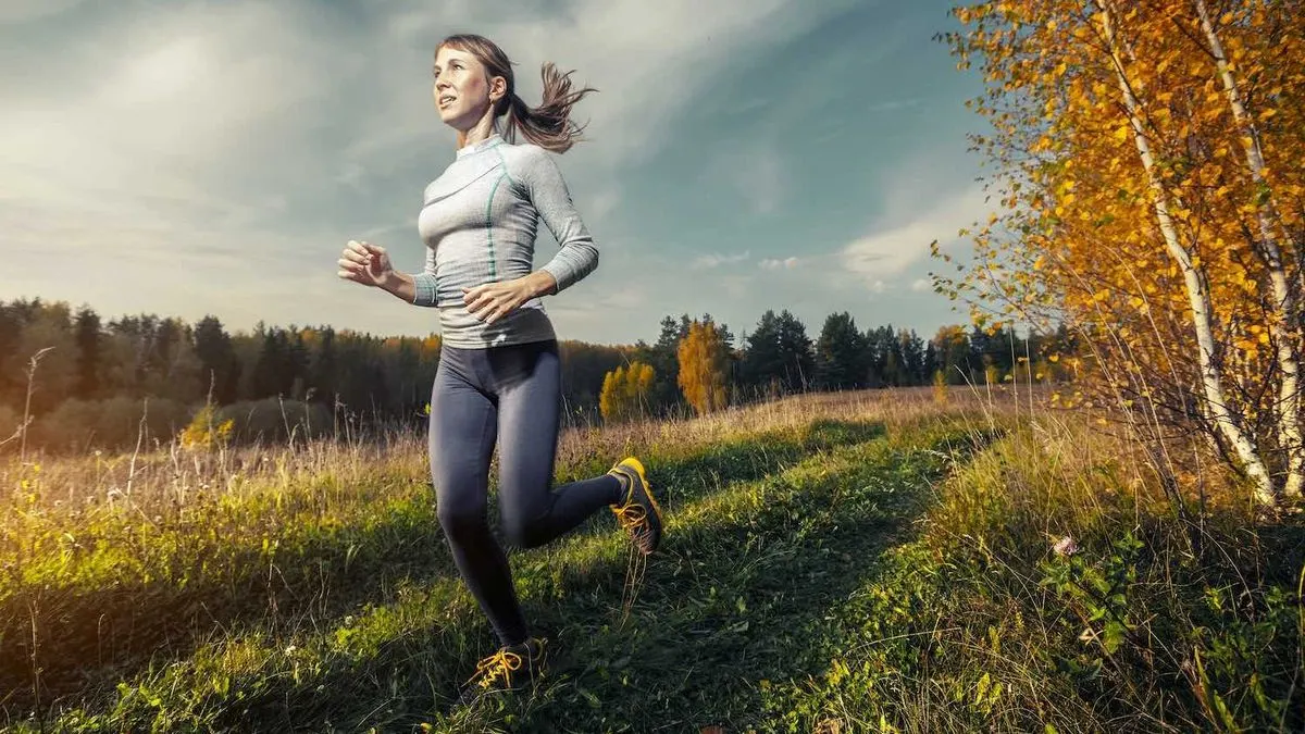 Frau joggt in Sportkleidung auf einem schmalen Pfad durch herbstliche Natur mit bunten Bäumen und Wiese.