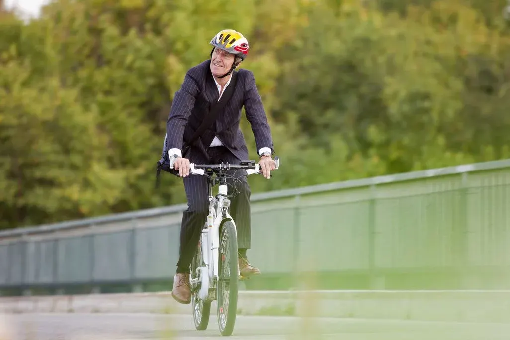 Mann im dunklen Nadelstreifenanzug und gelbem Helm fährt weißes E-Bike auf gepflastertem Weg. Grüne Bäume und Brückengeländer im Hintergrund.