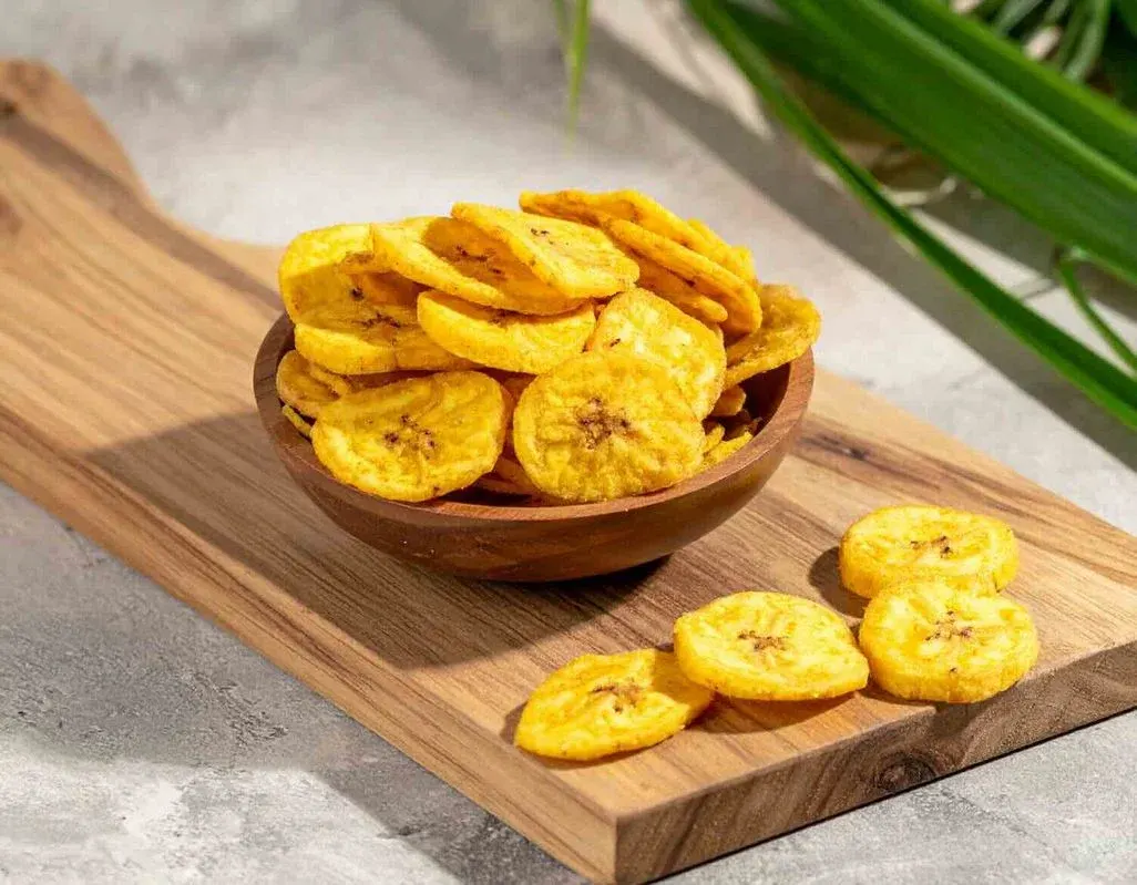 Goldgelbe knusprige Bananenchips in einer runden Holzschüssel, angerichtet auf einem hellen Holzschneidebrett. Einige Chips liegen zusätzlich lose auf dem Brett verteilt. Im Hintergrund rechts sind grüne Palmenblätter zu erkennen. Das Arrangement steht auf einer hellgrauen Steinoberfläche.
