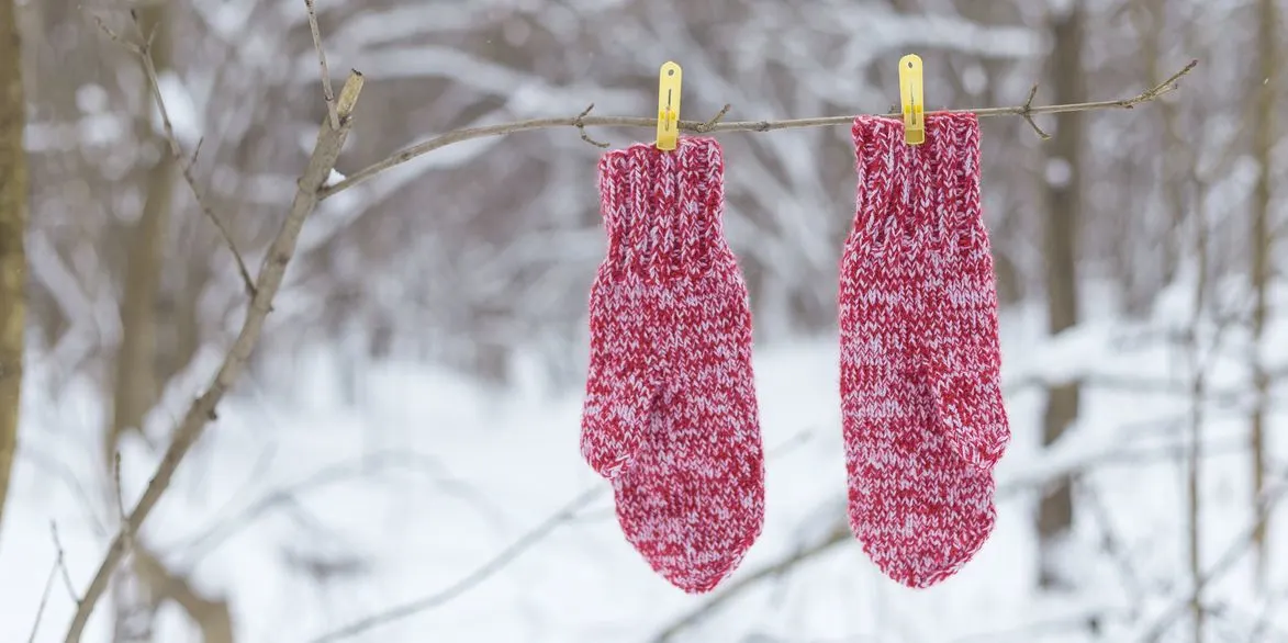 Zwei rot-weiße Strickhandschuhe hängen mit gelben Wäscheklammern an einem kahlen Ast vor verschneitem Winterwald.